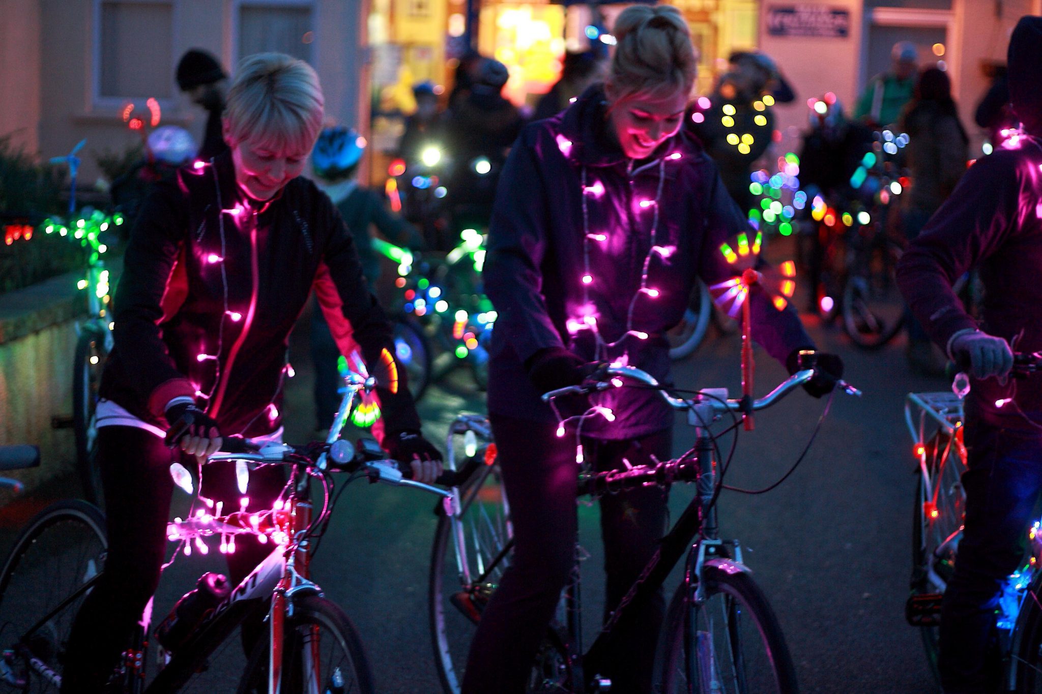 Image description: A group of cyclists are gathered together their bikes and themselves decorated with fairy lights of different colours, pink , green, blue etc. Two women at the front both smiling and looking at their bikes preparing to set off.